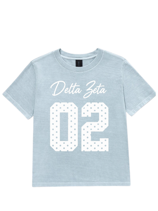 A light blue short-sleeve t-shirt with 'Delta Zeta' in white script and the number '02' in a white polka-dot block font on the front, displayed flat against a white background.