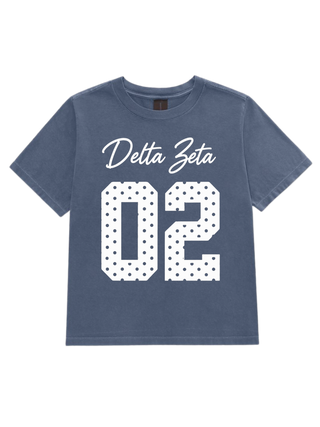 A short-sleeve crewneck t-shirt in a washed slate blue color, featuring a white graphic print on the front with the text 'Delta Zeta' in cursive and the number '02' filled with a polka dot pattern.